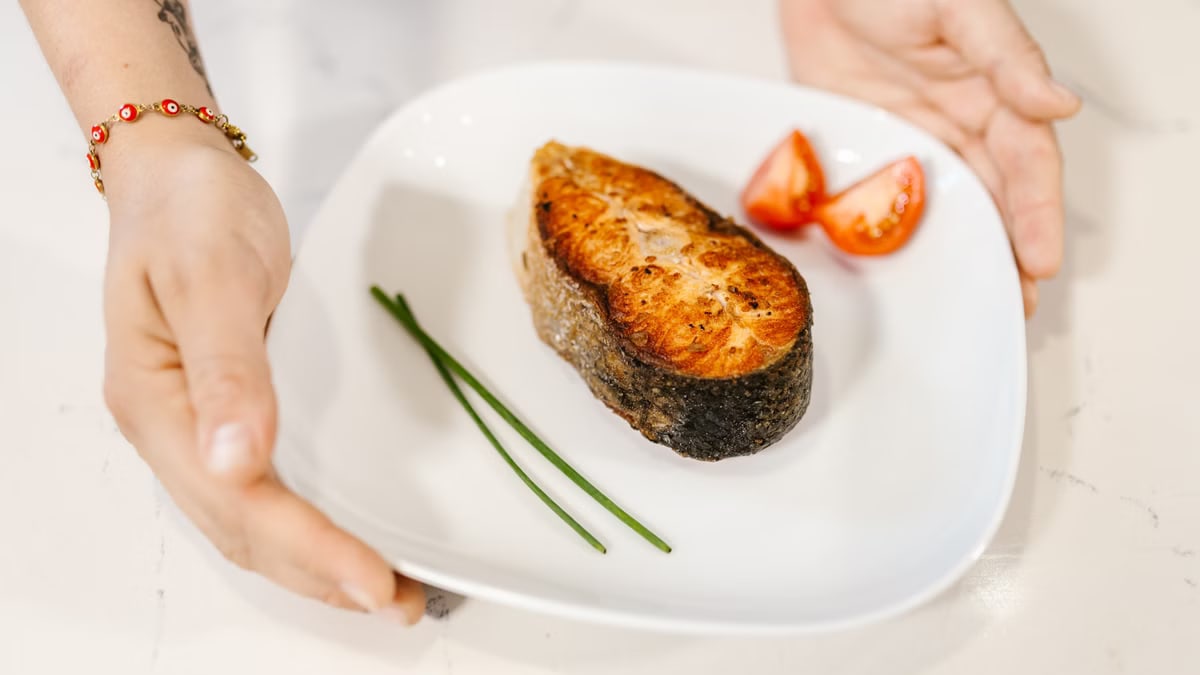 Close-up of grilled salmon served with tomatoes and chives on a plate, held by tattooed hands.